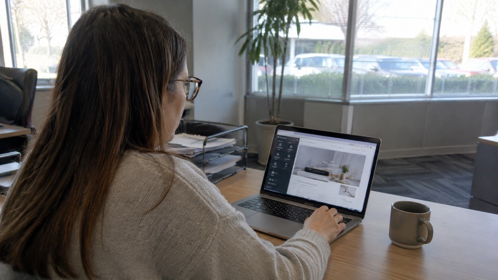 Femme à lunettes en pull beige travaillant sur une fiche produit e-commerce depuis un MacBook argenté