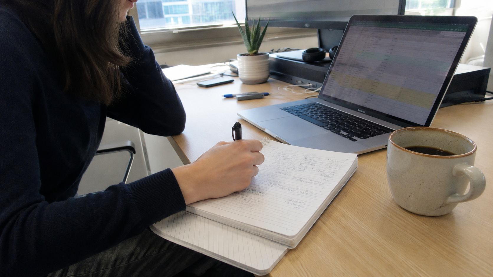 Femme en pull noir prenant des notes dans un carnet à spirale à côté d'un tableur ouvert pour arbitrer un budget d'outils SEO
