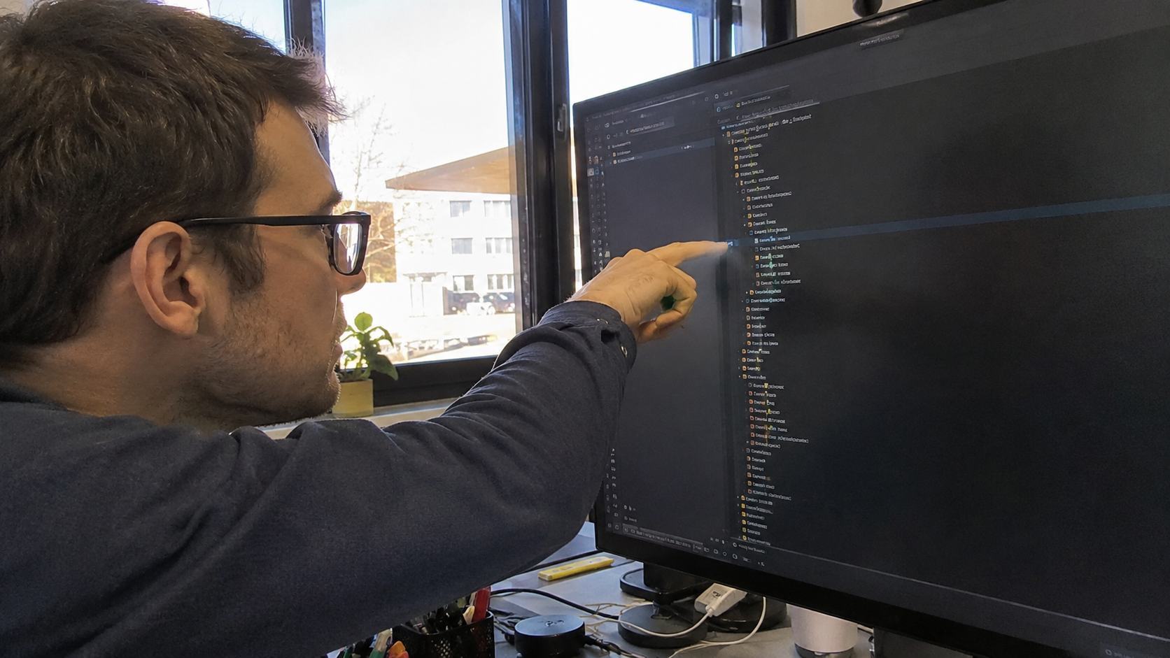 Consultant SEO en pull bleu marine désignant une arborescence de fichiers sur un écran de code, illustrant l'exploration des pages par Googlebot